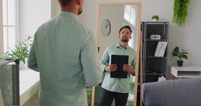 Man holds a clipboard, preparing for rehearsal and presentation, checking reflection in a mirror. Man rehearses presentation in front of mirror. Employee prepare for job conference at home