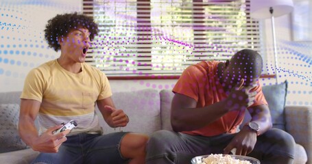 Reacting, two men in yellow orange shirts gaming on couch at home with white controller, popcorn