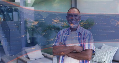 Standing mature man crossing arms facing camera in living room, in plaid shirt, glass reflections