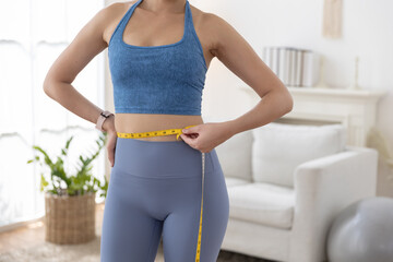 Close up of Asian woman in activewear measuring her waist with tape measure. Concept of weight loss, fitness progress, body transformation and healthy lifestyle. Great for health content.