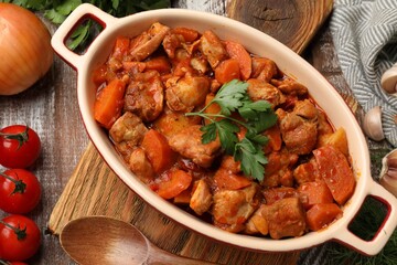 Delicious cooked stew in baking dish, fresh ingredients and spoon on wooden table, flat lay