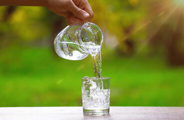 Man pouring water into glass from jug at wooden table against blurred green background, closeup