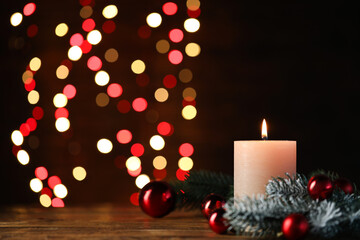 Burning candle, fir tree branches and baubles on wooden table against black background with blurred lights, closeup with space for text. Christmas greeting card