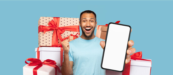 A man expresses joy while holding a smartphone in front of a colorful set of wrapped gifts. The background features a bright blue, enhancing the holiday spirit and celebration.