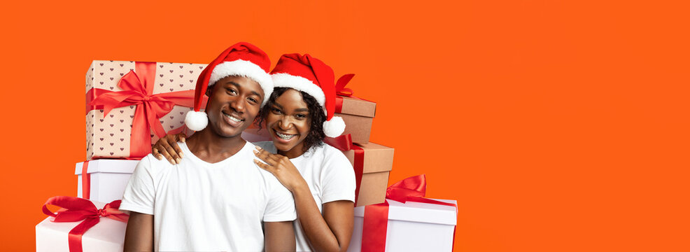 A joyful couple stands together wearing Santa hats, surrounded by colorful gift boxes. They share smiles while embracing the festive atmosphere and excitement of the holiday season.