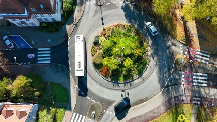Top-down aerial drone shot of a white bus and car navigating a city roundabout with a green center island and autumn trees. Traffic flow. - Powered by Adobe