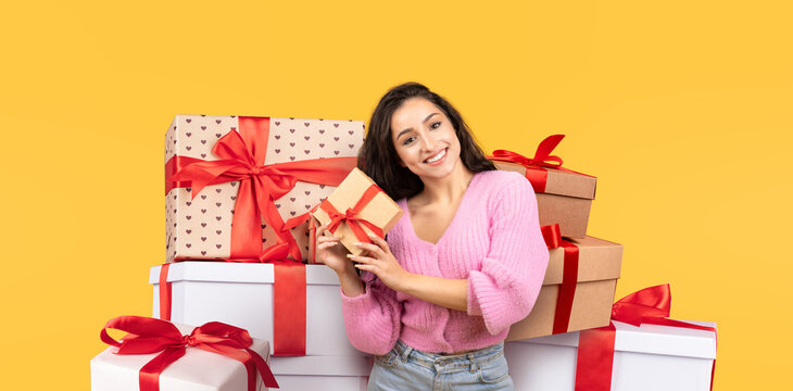 A cheerful woman stands among a pile of colorful gift boxes, holding a small present. The bright yellow background adds to the festive atmosphere, perfect for celebrations. - Powered by Adobe