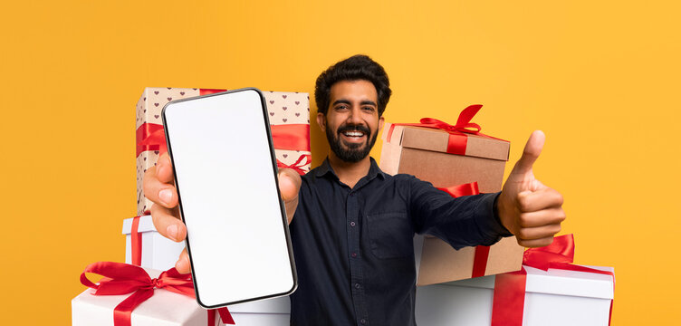A man smiles widely while holding a smartphone in one hand. Behind him, neatly stacked gift boxes are decorated with bows. The background is a vibrant yellow, adding to the festive feel.