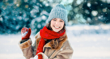 A young woman is smiling joyfully while holding a heart shape made of snow. She wears a warm winter outfit with a hat and scarf. Fluffy snow falls around her, creating a winter wonderland scene.