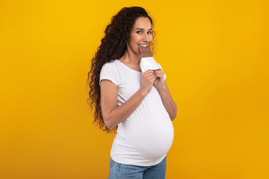 Pregnant lady stands in a studio with a bright yellow orange background. She bites a chocolate bar while smiling, capturing a joyful moment of sweet food cravings during pregnancy. - Powered by Adobe
