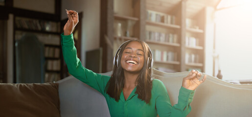 A young woman sits on a couch with headphones on, smiling and dancing joyfully. Sunlight streams...