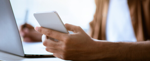 A person is focused on a smartphone while seated at a desk with a laptop. The scene captures a modern office environment with natural light, suggesting productivity and multitasking.