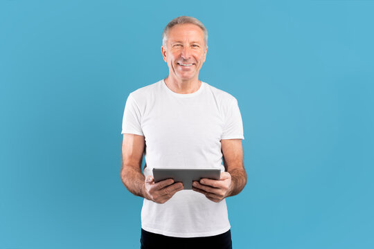 A cheerful mature man stands against a blue wall, holding a tablet and smiling. He appears comfortable and confident, engaged in using the device, showcasing a positive attitude towards technology. - Powered by Adobe