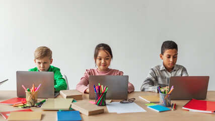 Three multiracial school kids are focused on their laptops, learning online at desks in a bright classroom. They browse the internet for educational activities during class time.