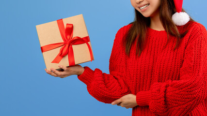 A young woman wearing a red sweater and a Santa hat is smiling while she holds a gift box with a red ribbon. The background is bright blue, creating a cheerful holiday atmosphere.