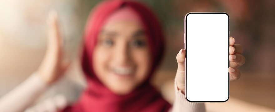 A young woman in a red hijab joyfully displays a smartphone with no content visible. She is smiling and giving a thumbs-up, highlighting a casual moment in a warm environment.
