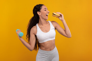 A happy woman stands against a vibrant yellow background, holding a tasty burger in one hand and a dumbbell in the other. She embodies the balance of an unhealthy diet and a healthy lifestyle.