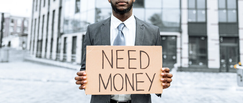 A man dressed in a suit stands outdoors in a city, holding a cardboard sign that reads need money. The background features modern buildings and a clear sky.