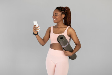 A lovely young African American woman in sports clothing smiles as she holds a fitness mat and checks her mobile phone. She is likely getting ready for a yoga or pilates workout in a studio.