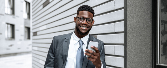 A man dressed in a suit with glasses smiles while holding a smartphone. He stands against a sleek gray wall in an urban setting, looking relaxed and engaged in his activity.