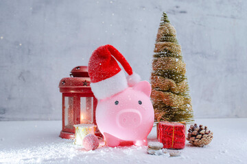 Pink piggy bank wearing Santa hat with Christmas tree, red lantern, gift boxes and snow. Holiday concept for Christmas savings and festive finance planning.