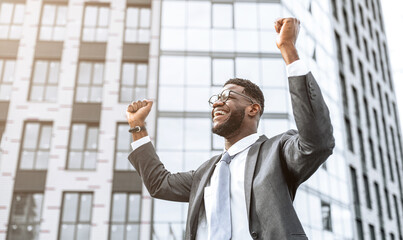 A man in a suit stands outside a contemporary office building. He smiles and raises his arms in triumph, celebrating a recent success or accomplishment. © Prostock-studio
