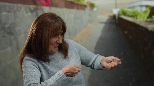 Hispanic senior woman playfully mimics pulling a rope on a city street, showcasing joyful expression in an outdoor urban setting with stone walls and vibrant atmosphere.