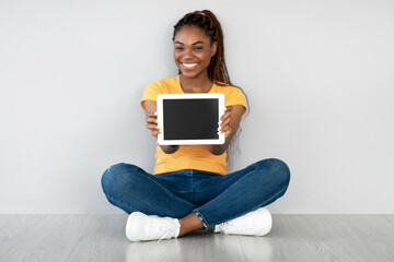 A beautiful black woman sits on the floor against a grey wall, smiling while holding up a tablet computer with an empty screen. She is demonstrating a template for a website or application.