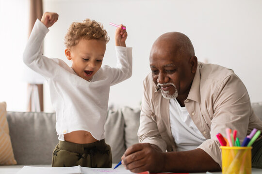 Two family members enjoy quality time together as they sketch at home. The grandfather carefully draws while the excited grandson celebrates with joy, creating a fun and artistic atmosphere. - Powered by Adobe