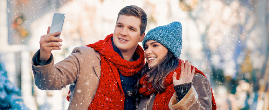 A young couple enjoys a snowy winter day while taking a selfie together. They are dressed warmly, with cozy coats and colorful scarves. - Powered by Adobe