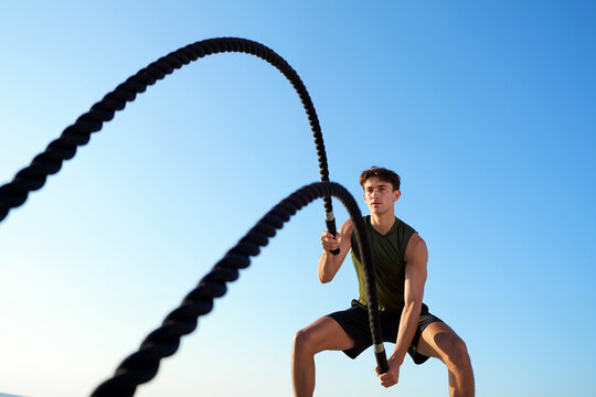 A dedicated young man engages in an intense workout using battle ropes in an outdoor setting under a bright blue sky during daylight. Generative AI.