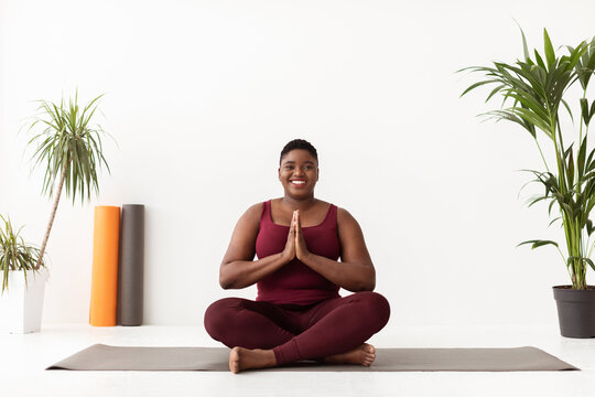 Cheerful plus size black woman is sitting in lotus position on a fitness mat in a yoga studio. She wears sportswear and smiles at the camera, exuding positivity and relaxation.