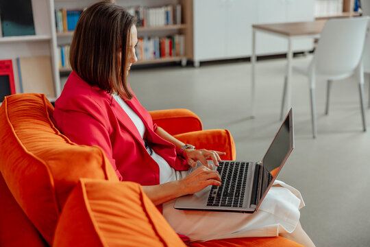 woman working on laptop sitting on bright orange sofa in modern interior concept of remote work education business coaching and digital communication