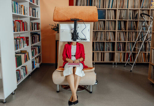 Unrecognizable woman sits in a modern coworking space. She wears a red blazer and white dress, typing on a laptop. Bookshelves are in the background.