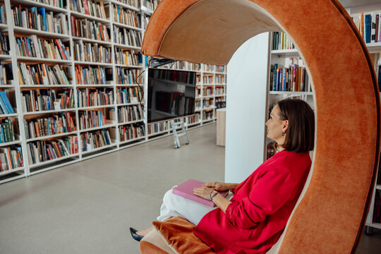 A young Hispanic woman with brown hair sits in a modern orange chair, working on a laptop in a coworking space filled with bookshelves. - Powered by Adobe