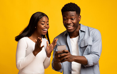 Two young adults are expressing joy as they look at a smartphone together. They share excitement while standing in front of a bright yellow backdrop, creating a vibrant and cheerful atmosphere.