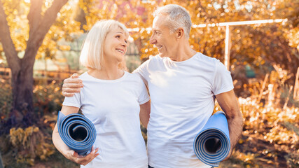 In a vibrant outdoor setting during autumn, a senior couple stands together holding yoga mats, smiling and enjoying each other's company. The warm colors of the season enhance the cheerful atmosphere.