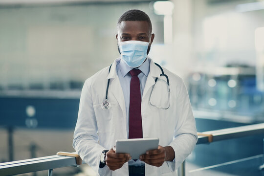 African american young man doctor in workwear and face mask with stethoscope posing with modern digital tablet at modern clinic, copy space. Online appointment, remote healthcare, telemedicine service