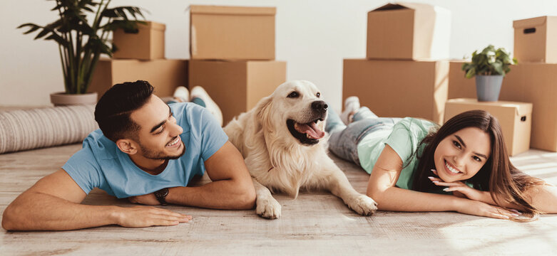 A young couple lies on the floor with their golden retriever in a newly moved-in home. They appear joyful, surrounded by cardboard boxes and a houseplant.