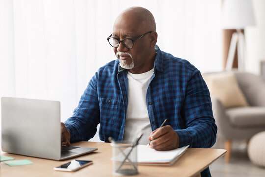 A senior black man sits at his office desk, working on a laptop and taking notes. He wears glasses and shows concentration as he writes in a notebook. This reflects entrepreneurship in a mature age.