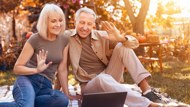 A happy couple sits on a blanket in a park during autumn, using a laptop to connect with friends. They wave and smile, surrounded by colorful leaves and pumpkin decorations.