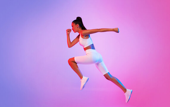 A sporty lady showcases her athleticism by running in mid air, wearing white fitwear against a striking pink and blue neon background.