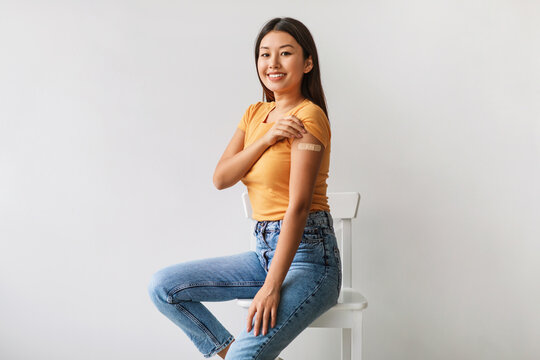 A cheerful Asian woman sits on a chair, revealing her shoulder with an adhesive bandage after receiving the covid-19 vaccine. She smiles happily, signifying health protection against viral diseases.