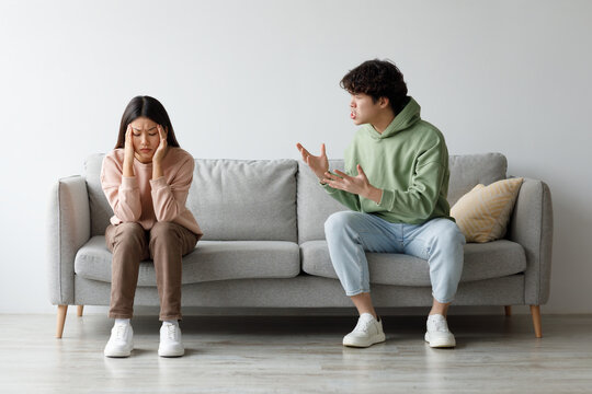 A millennial Asian couple sits on a couch at home, engaged in a heated argument. The man is shouting, while the woman appears upset, covering her face with her hands.