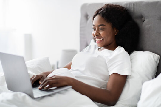Young african american woman types on her laptop in bed. She is smiling and wearing pajamas as she enjoys her remote freelance work. Bright, cozy space enhances her productive moments.