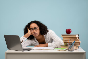 Tired black female student studies at desk surrounded by books and a laptop. She rests her head on her hand, showing signs of exhaustion, with a blue background emphasizing her struggle.