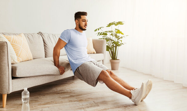 A man performs a tricep dip exercise using his couch as support in a bright living room. He stays focused while enhancing his fitness routine at home.