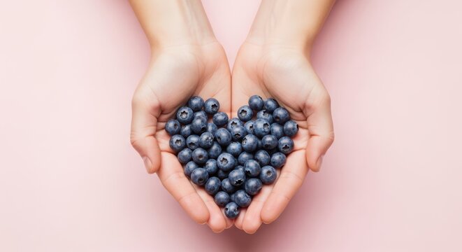 Hands holding a heart shape filled with fresh blueberries against a soft pink background, symbolizing love for healthy eating and natural goodness