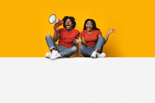 A couple sits on the floor, both wearing red shirts and jeans, expressing excitement. One holds a megaphone while the other waves their hands joyfully in a vibrant, cheerful setting. - Powered by Adobe