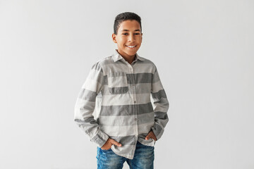 Naklejka premium A cheerful African American preteen boy stands with a friendly smile, hands in pockets, wearing casual clothes in a gray studio setting. His joyful appearance suggests confidence and happiness.